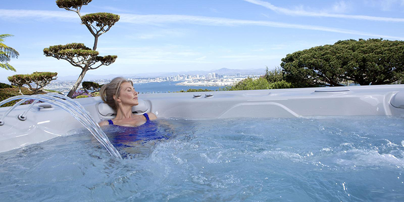 Woman relaxing in a luxurious outdoor hot tub with cascading water jets, overlooking a stunning cityscape and ocean view under a clear blue sky.