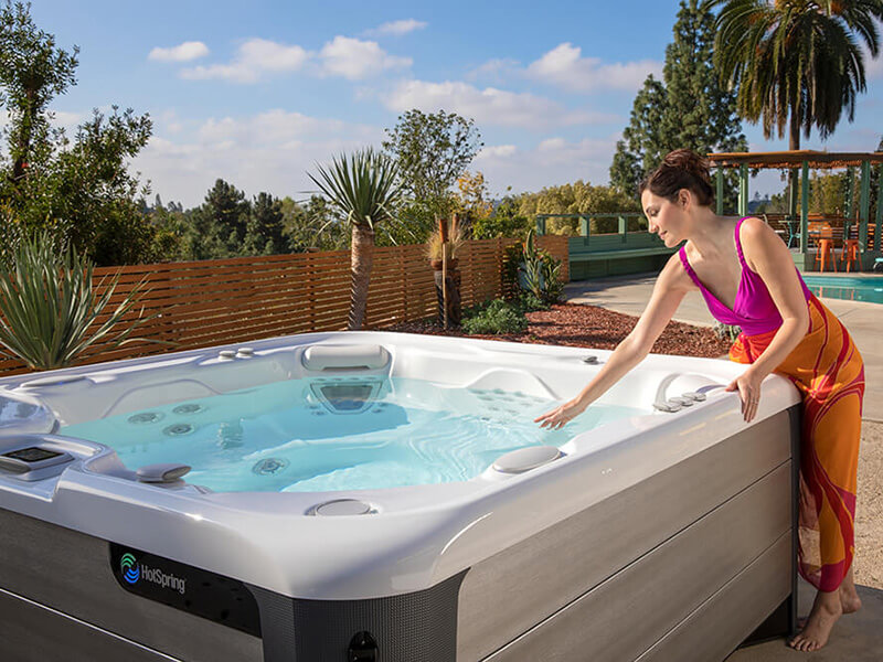 A woman gently touching the water of her hot spring saltwater spa in an outdoor setting, with a swimming pool visible in the background.