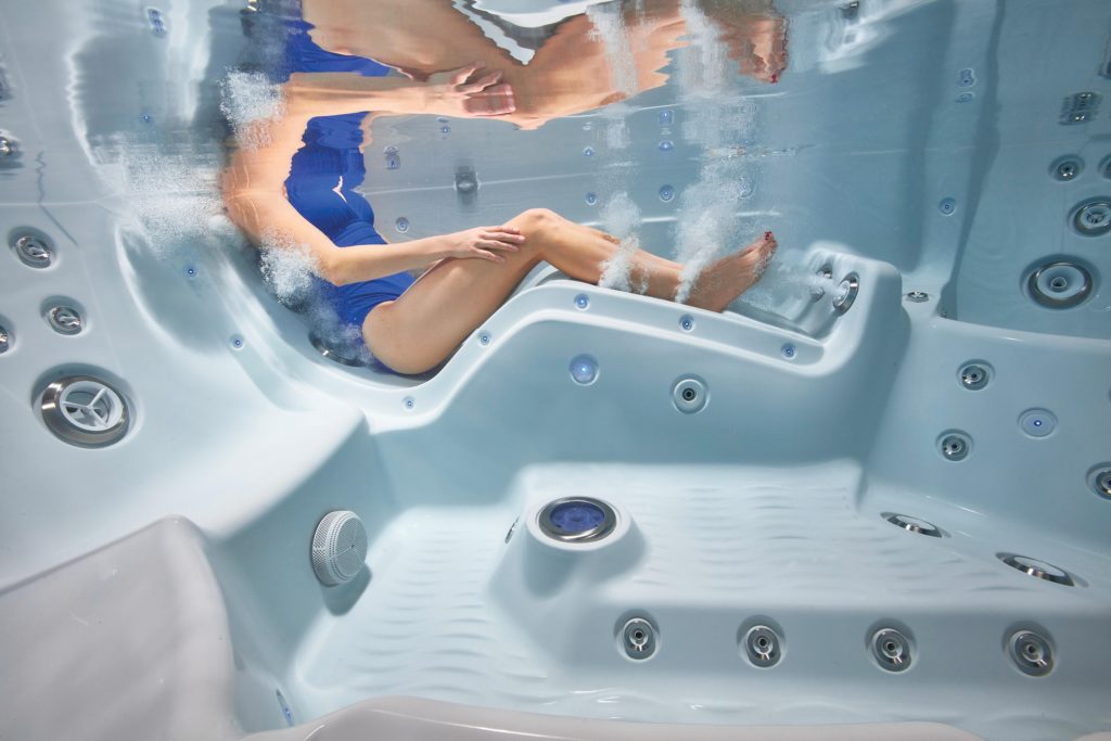 Underwater view of a woman in a blue swimsuit reclining in a hot tub, with powerful hydrotherapy jets massaging her legs and back.