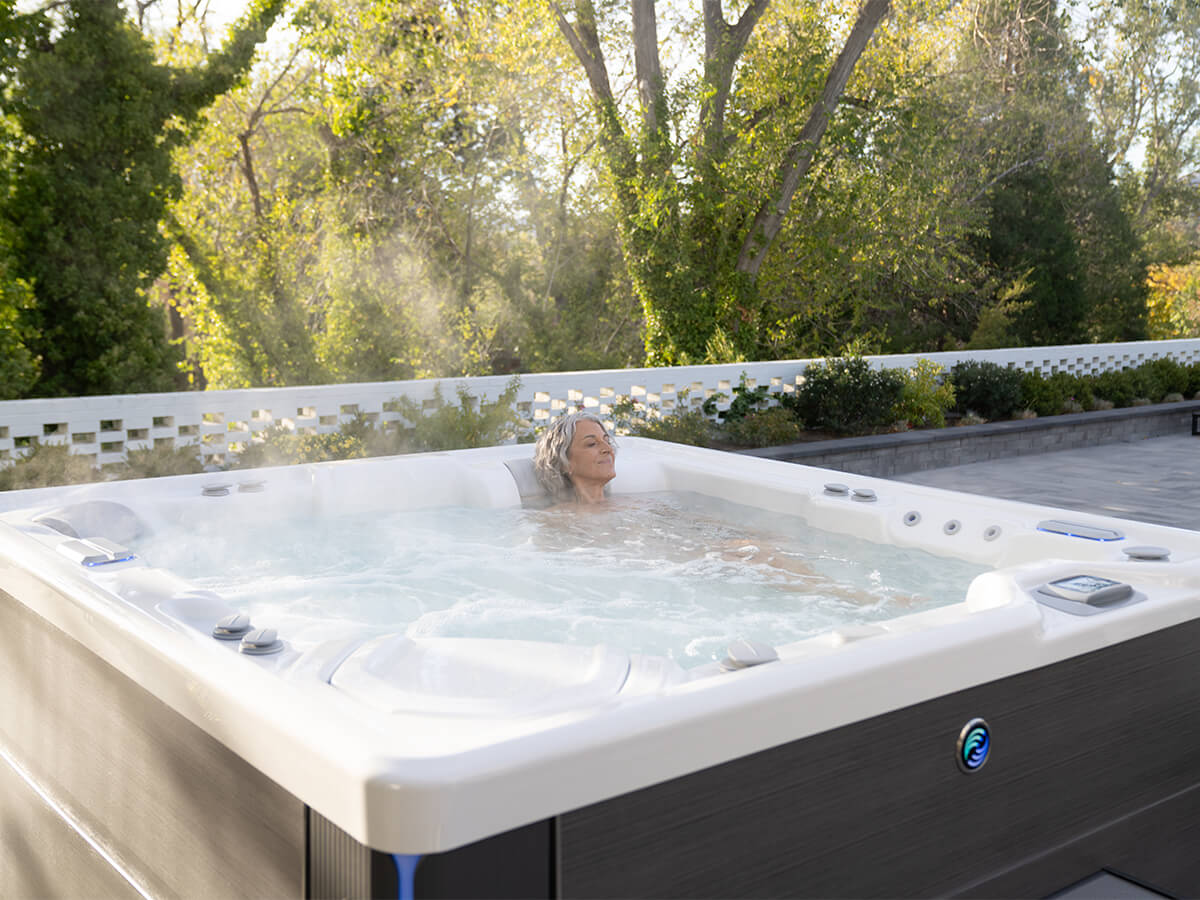 Hot Spring Spas Hot Tub: The Perfect Addition to Your Backyard Design. A woman is enjoying a soak in her Hot Spring Spas hot tub in an outdoor backyard setting. The hot tub is surrounded by lush greenery. The woman is relaxed and content, enjoying the peace and quiet of her backyard.