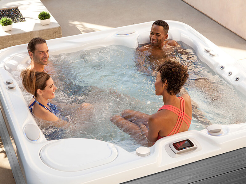 A group of four friends enjoying a relaxing soak in a Hot Spring saltwater hot tub on a sunny afternoon. They are laughing and unwinding in the warm, soothing water, surrounded by a peaceful outdoor setting.