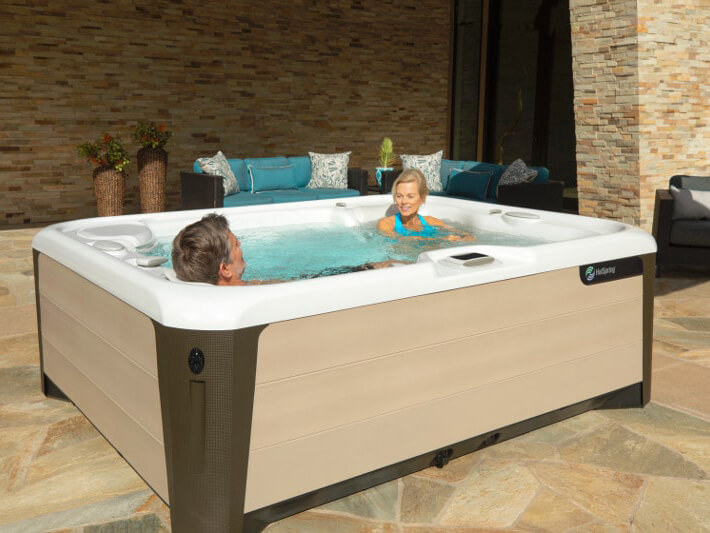 Smiling older couple relaxing in a HotSpring spa on a stylish patio, surrounded by plush outdoor seating, decorative pillows, and a stone accent wall.