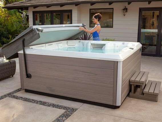 Woman lifting and inspecting the insulated cover of a modern outdoor hot tub on a patio, with a house and greenery in the background.