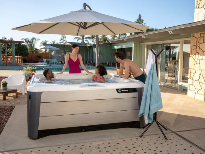 Group of friends and family enjoying a conversation in a HotSpring spa under a large patio umbrella, with a modern home, pool, and lush landscaping in the background.