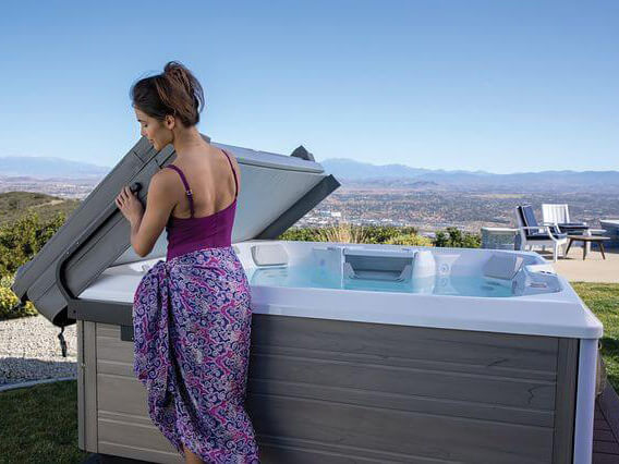 Woman in a purple swimsuit and patterned sarong lifting the cover of an outdoor hot tub, with a scenic mountain and city view in the background.
