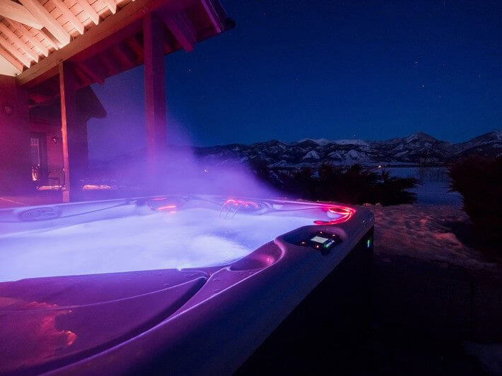 Steaming outdoor hot tub with glowing LED lights on a snowy patio, overlooking a breathtaking mountain range under a starry night sky.