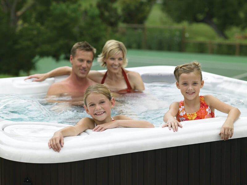 Smiling family of four enjoying a hot tub, with two young girls leaning on the edge while their parents relax in the bubbling water, surrounded by greenery.