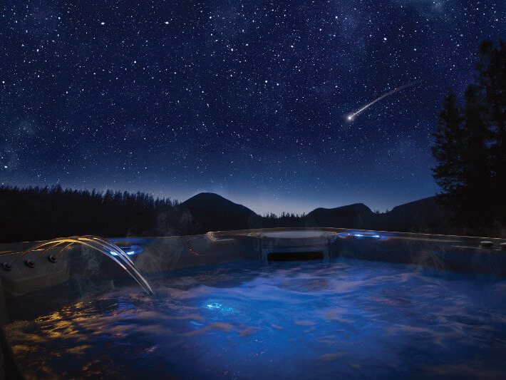 Outdoor hot tub illuminated with blue LED lights, steaming under a star-filled night sky with a shooting star, surrounded by mountains and trees.