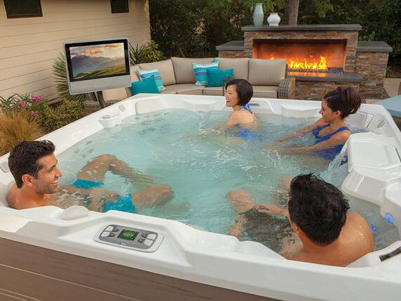 A group of friends relaxes in a luxurious Hot Spring Spa outdoor hot tub. The hot tub area is decked out with premium accessories: a large flat-screen TV mounted nearby and high-quality speakers strategically placed for optimal sound.