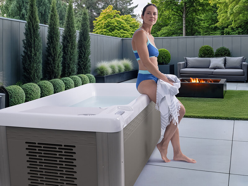 Woman sitting beside a Hot Spring cold plunge tub in a modern backyard with a fire feature.