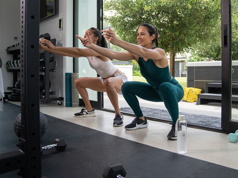 Two women doing squats together in a home gym before a hot and cold contrast therapy session.